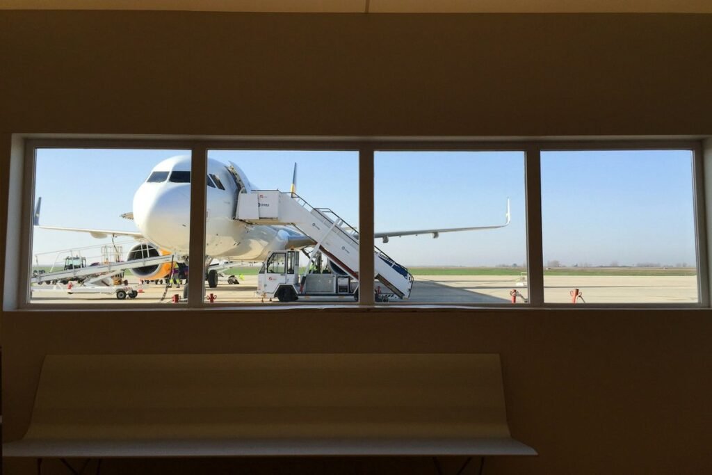 A white passenger jet parked at the gate with boarding stairs, seen through a row of windows from a quiet departure lounge with an empty bench