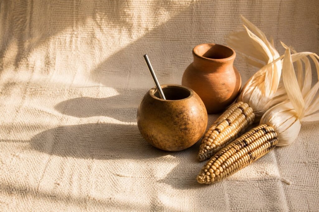 A calabash mate gourd with a metal straw, a terracotta clay pot and dried maize on a cream woven cloth