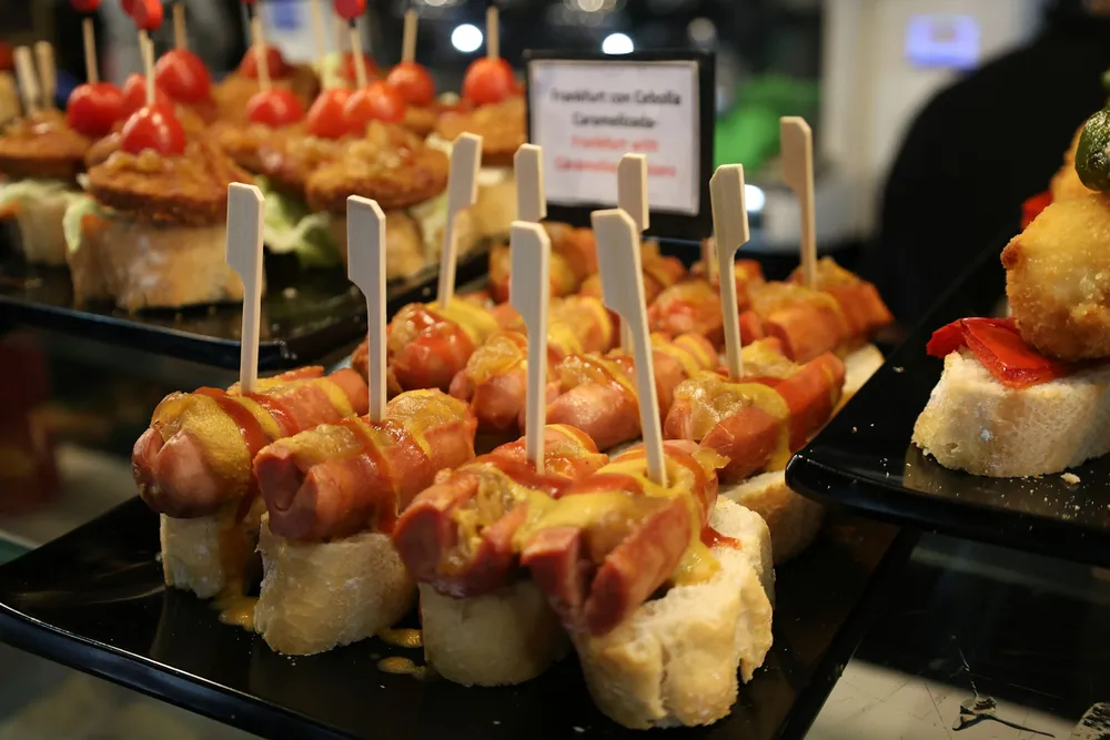 Pintxos of sausage and mustard on bread, lined up on a bar