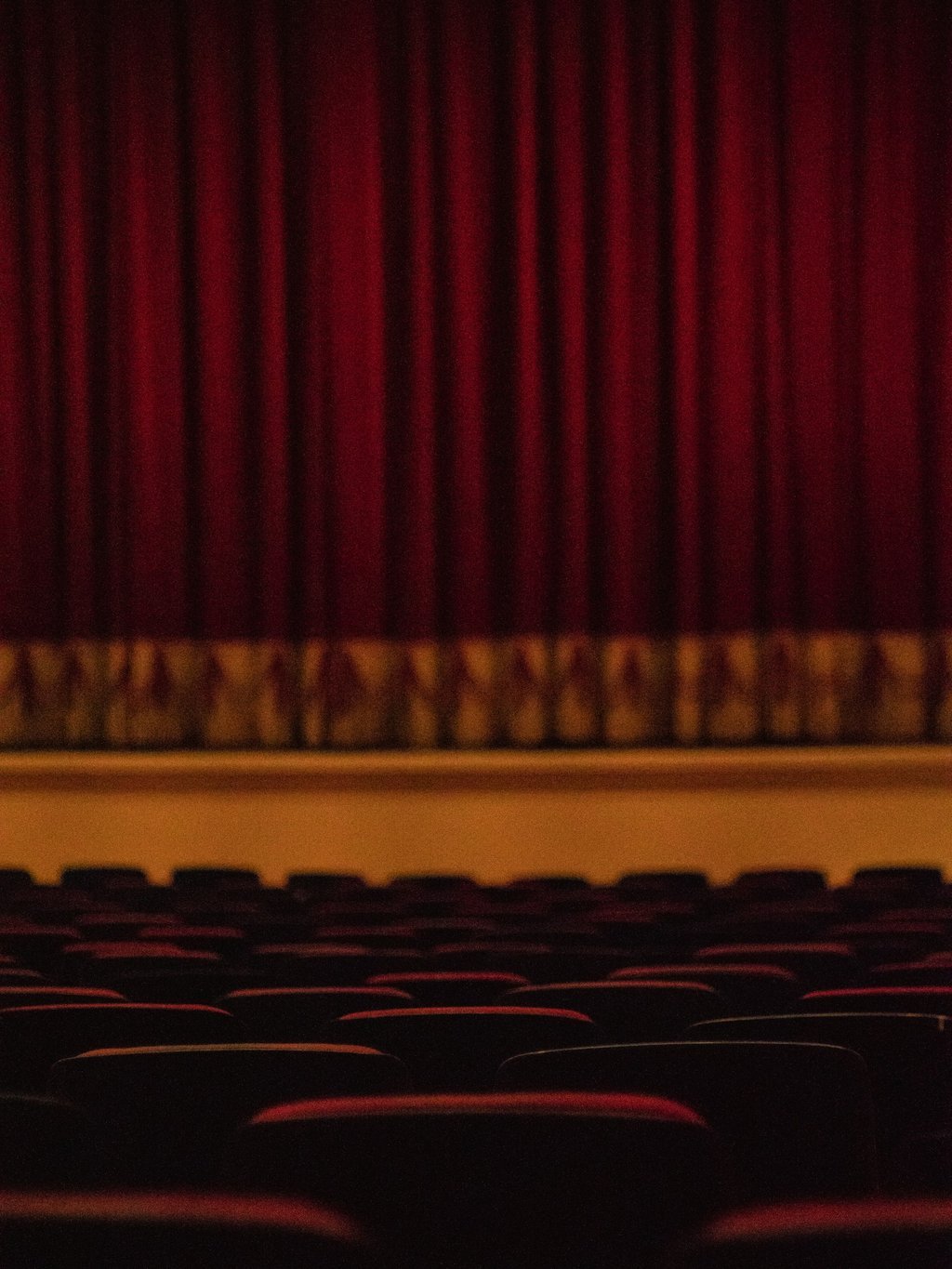 An empty cinema with a red velvet curtain over the stage and dark rows of seats in front