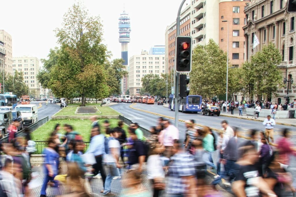 A busy crossing in central Santiago de Chile, with walkers blurred by motion, trees, buses and the Torre Entel rising behind the avenue